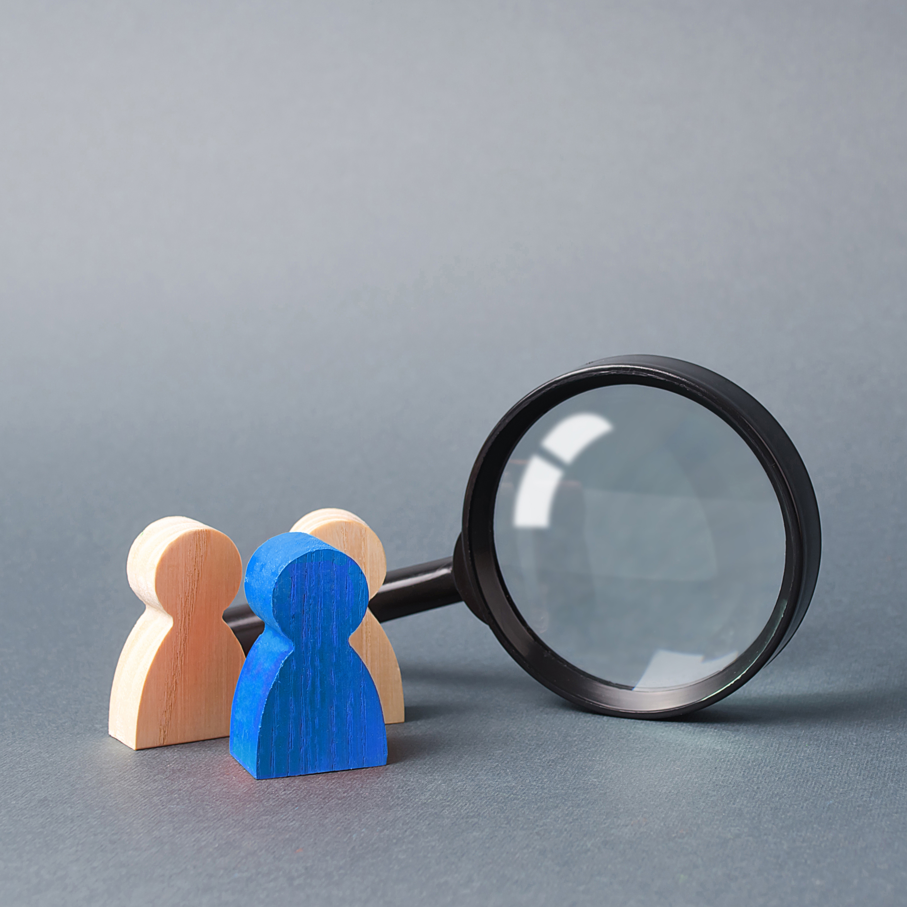 Three wooden human figure stands near a magnifying glass on a gray background.